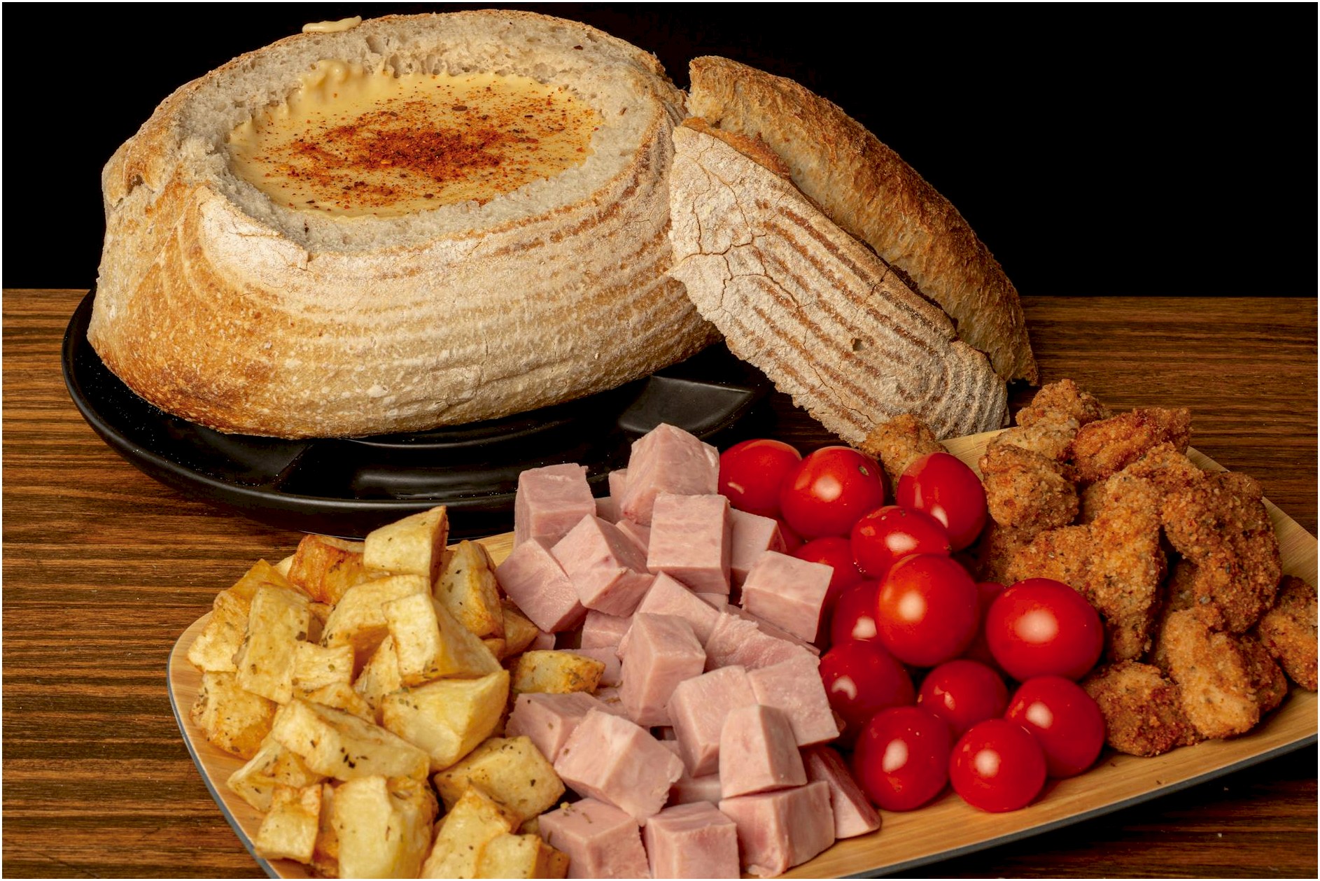 Delicious rustic bread bowl filled with cheese, surrounded by deli meats and fresh vegetables.