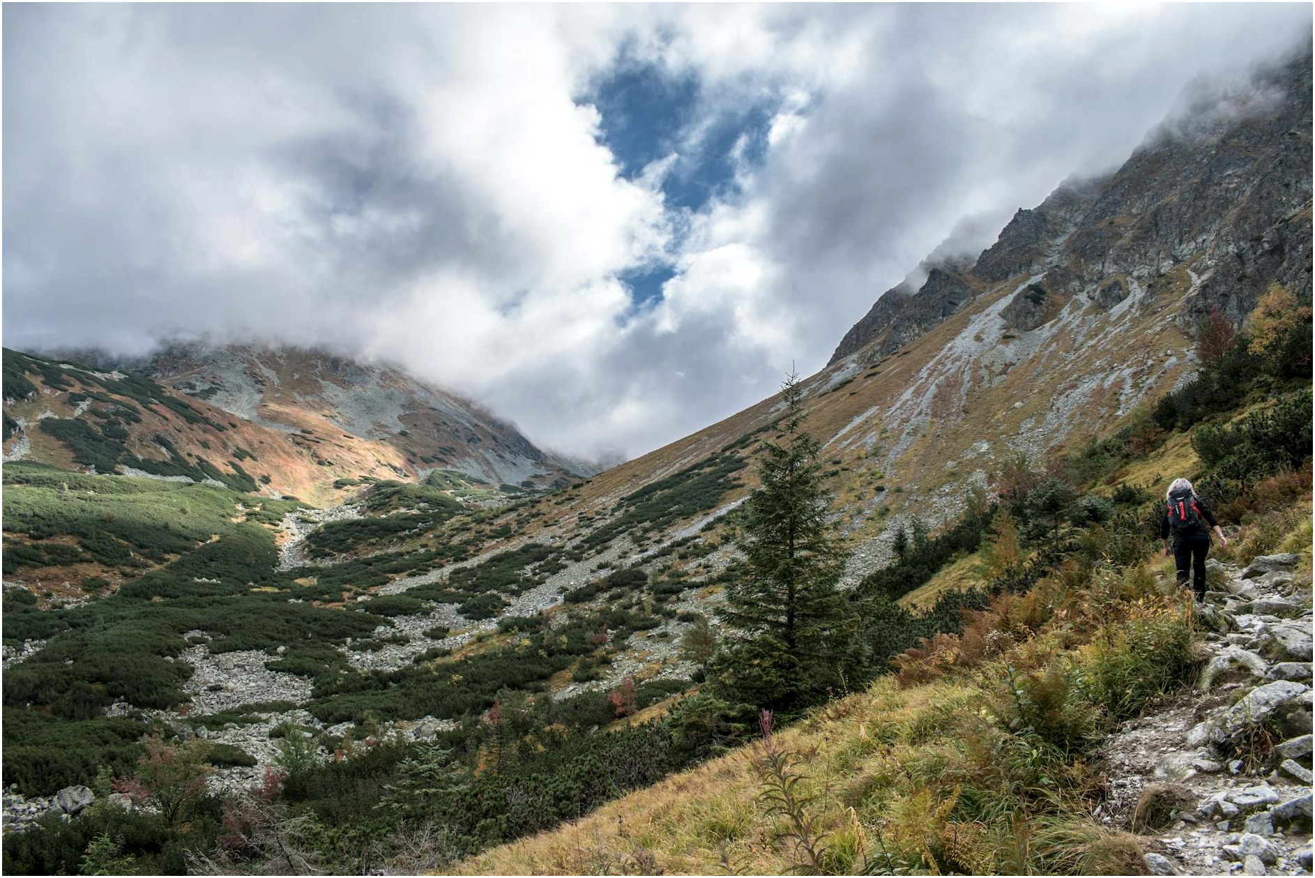 Woman hiking on a scenic rocky trail in mountainous terrain under a cloudy sky.